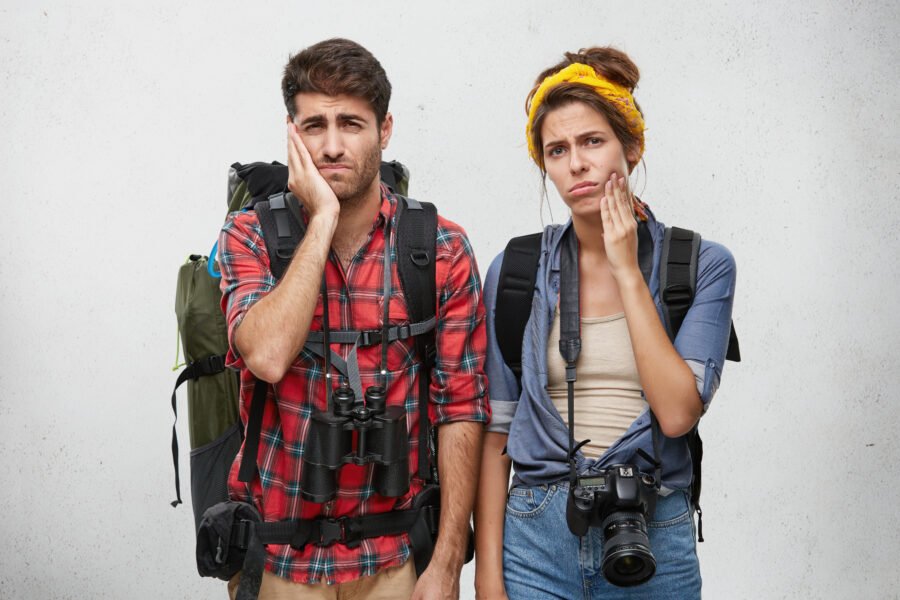 A young tourist couple with backpacks holding their hands to their cheeks, as if suffering from a toothache.