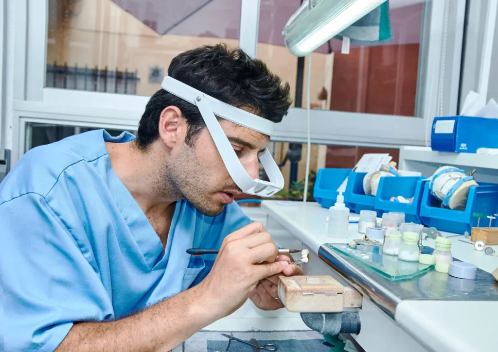 Dental technician wearing magnifying glasses, layering porcelain by hand onto a crown in a dental lab.