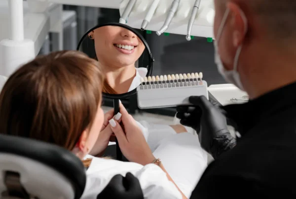 Patient smiling into a hand mirror while her dentist holds up a shade guide to choose the colour of her veneers.