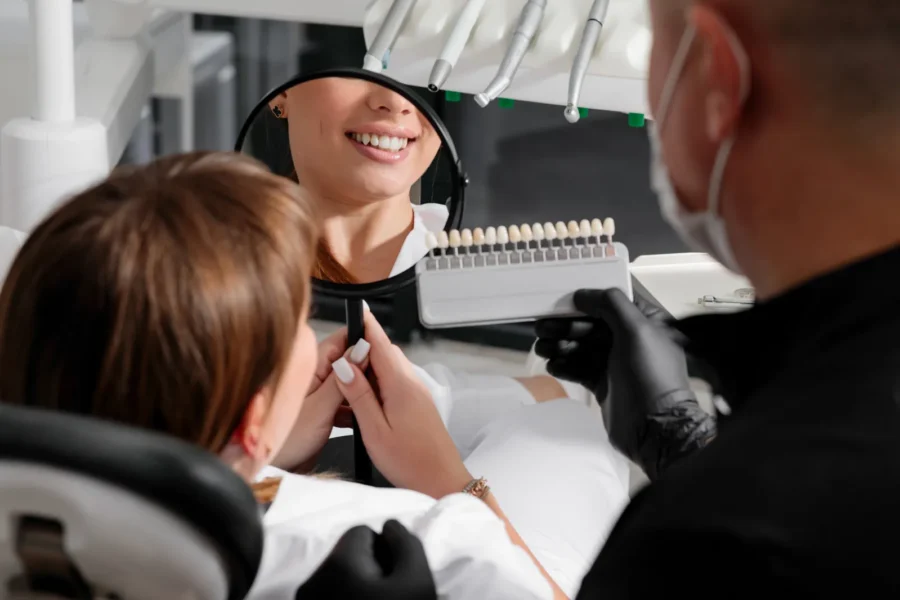 Patient smiling into a hand mirror while her dentist holds up a shade guide to choose the colour of her veneers.